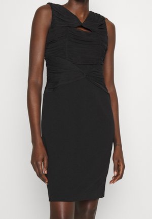 Ärmelloses schwarzes knielanges Kleid mit gerafftem Oberteil und gedrehtem Detail an der Taille, getragen von einer stehenden Person vor einfarbigem Hintergrund.