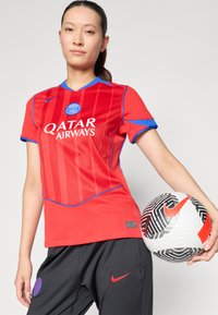 Röd Nike-tröja med blå accenter och vertikala ränder, med "Qatar Airways" på framsidan; tillsammans med svarta byxor och en mönstrad fotboll.