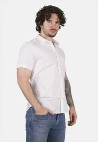 Camicetta bianca a maniche corte, design con bottoni, texture liscia, con colletto standard. Indossata con jeans blu, mostrando un look casual.