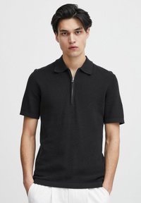 Polo shirt scurt negru cu un textil moale și ribbed, având un sistem de închidere cu fermoar și un design clasic al gulerului.