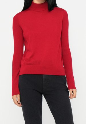 Kvinde iført en tætsiddende rød rullekravesweater og sorte bukser, stående mod en ensfarvet lys baggrund.
