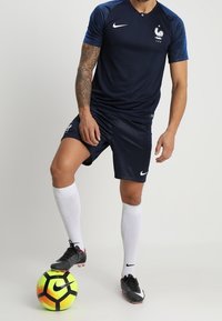 Muž v dresu francouzské fotbalové reprezentace s logy Nike, v bílých ponožkách a kopačkách, stojící s nohou na žlutočerném fotbalovém míči Nike.