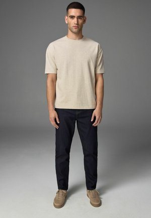 Beige katoenen t-shirt met korte mouwen, gecombineerd met donkerblauwe jeans en tan sneakers. Man staat tegen een grijze achtergrond.
