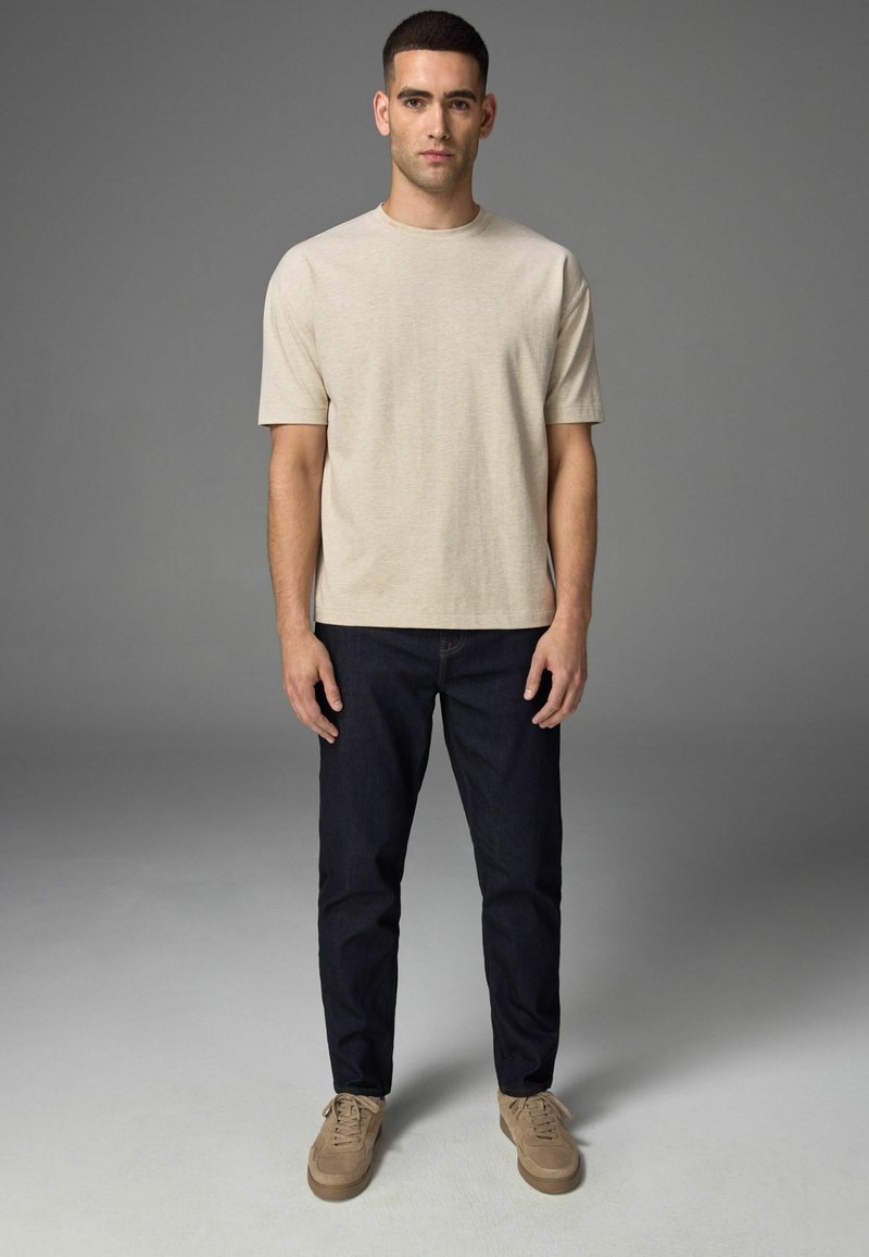 Camiseta de algodón beige de mangas cortas, combinada con jeans azul oscuro y zapatillas marrones. Hombre de pie contra un fondo gris.