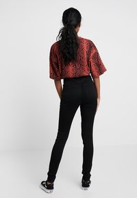 Žena s dlouhými černými vlasy v culíku, oblečená v červeném topu se vzorem hadí kůže, černých skinny džínách a černých teniskách, stojí čelem k odvrácené straně.