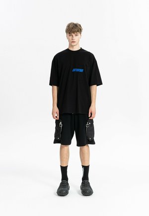 Joven de pie con una camiseta negra de gran tamaño con logo azul, pantalones cortos cargo negros con cremalleras, calcetines negros y zapatos negros con textura.