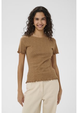 Uśmiechnięta kobieta o kręconych włosach, ubrana w dopasowany brązowy sweter z krótkim rękawem i jasnobeżowe spodnie na jednolitym tle.