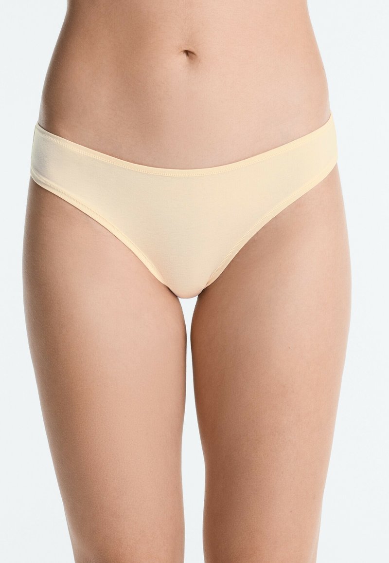Femme portant un sous-vêtement style bikini en coton beige, montrée du milieu du torse aux cuisses supérieures sur un fond uni.