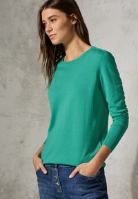 Grüner Rundhalsausschnitt-Pullover aus weichem Material, mit langen Ärmeln und gerippten Bündchen, kombiniert mit blauer Jeans mit Knopfverschluss.