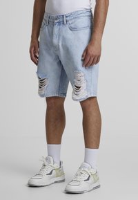 Lyseblå denimshorts med slidte knæhuller, standardtalje og oprullede kanter, parret med hvide og lysegrønne sneakers.