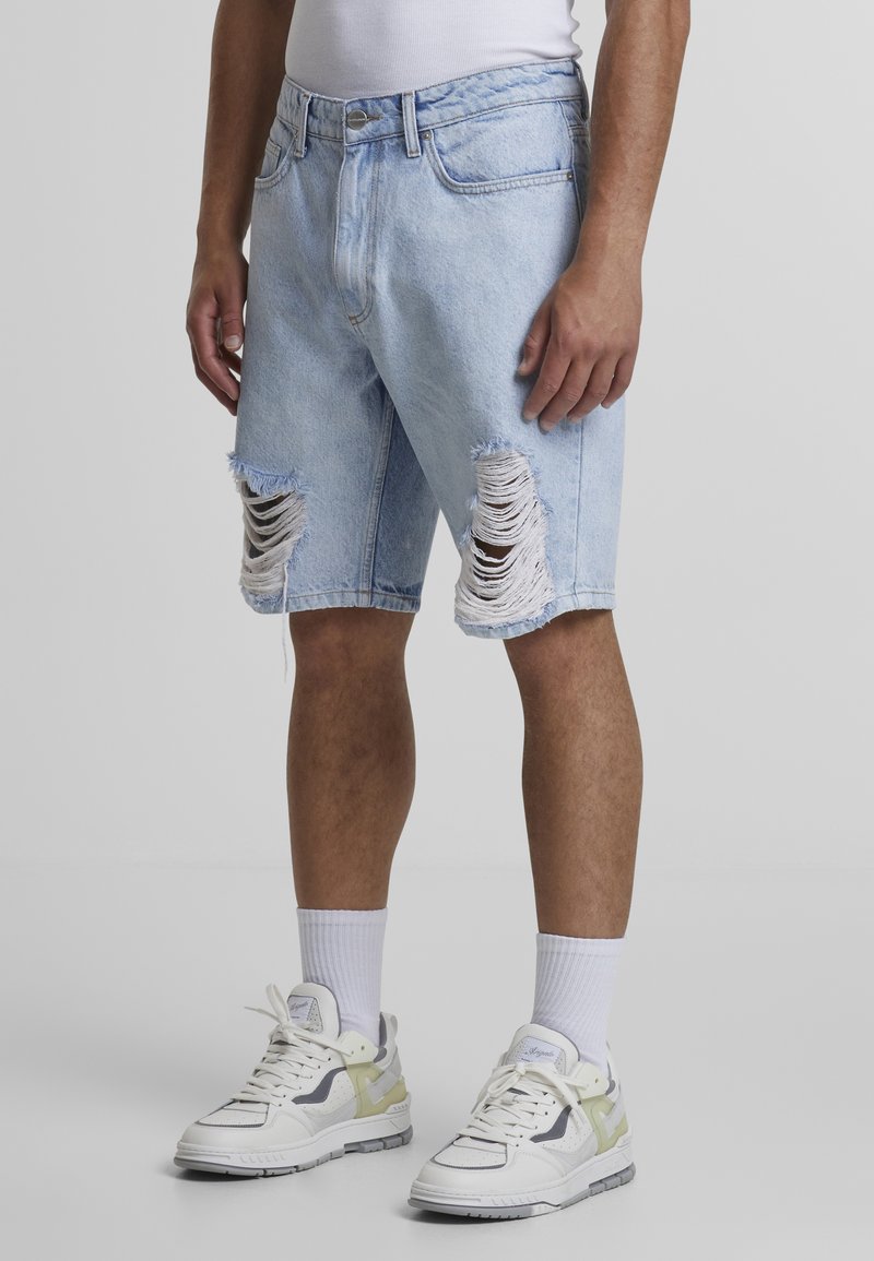 Lyseblå denimshorts med slidte knæhuller, standardtalje og oprullede kanter, parret med hvide og lysegrønne sneakers.