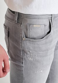 Jeans de denim gris con una ligera textura desgastada, bolsillos frontales y un acento de logo metálico. Vista en primer plano mostrando una mano en el costado.