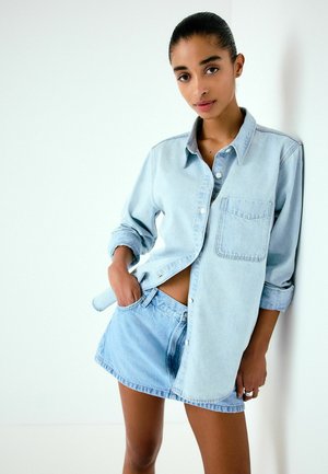 Ung kvinde lænende sig op ad væggen, iført lyseblå denimskjorte med optrukne ærmer og matchende denim-mininederdel, hånd i nederdelslomme.