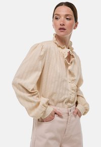 Blouse crème avec des rayures texturées et de petits pois, dotée d'un col à volants et d'une patte de boutonnage, associée à un pantalon rose clair.