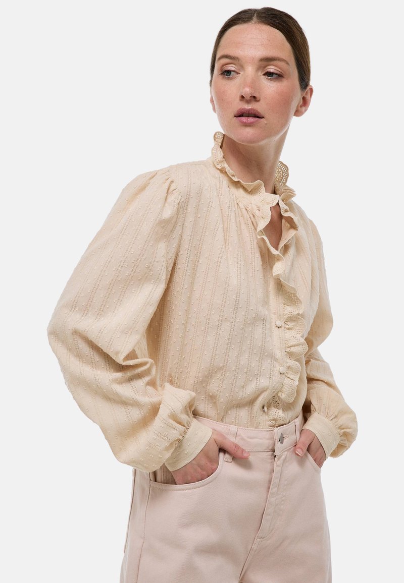 Blouse crème avec des rayures texturées et de petits pois, dotée d'un col à volants et d'une patte de boutonnage, associée à un pantalon rose clair.