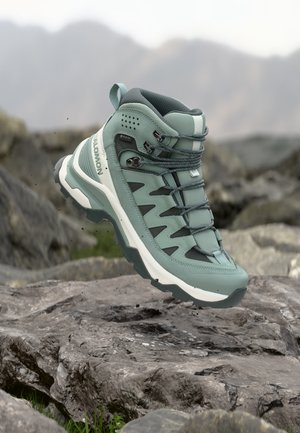 Light green hiking boot with rugged sole and Gore-Tex label hovering above rocky terrain, mountainous background blurred.