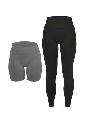 Frau trägt hoch taillierte schwarze Leggings und weiße Sneakers, steht mit den Armen entspannt an den Seiten vor einem schlichten Hintergrund.
