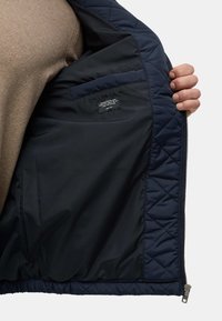 Gestepptes Innenfutter der dunkelblauen Jacke mit glattem Futter, seitlicher Tasche und sichtbarem Etikett. Reißverschlussöffnung an der Vorderseite. Strukturiertes Gewebe.