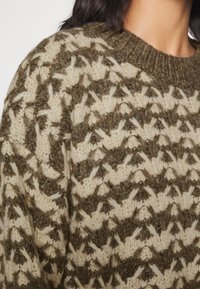 Detailní záběr osoby v silném, strukturovaném pleteném svetru se zeleno-krémovým geometrickým vzorem a žebrovaným kulatým výstřihem.