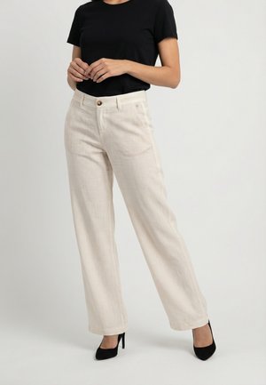 Donna che indossa pantaloni beige a vita alta e gamba larga, top nero a maniche corte e scarpe nere con tacco alto, in piedi davanti a uno sfondo semplice.