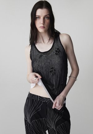 Frau mit langen dunklen Haaren, die ein durchscheinendes schwarzes Tanktop über einem weißen Hemd und schwarze Hosen mit weißen abstrakten Linien vor einem einfarbigen Hintergrund trägt.