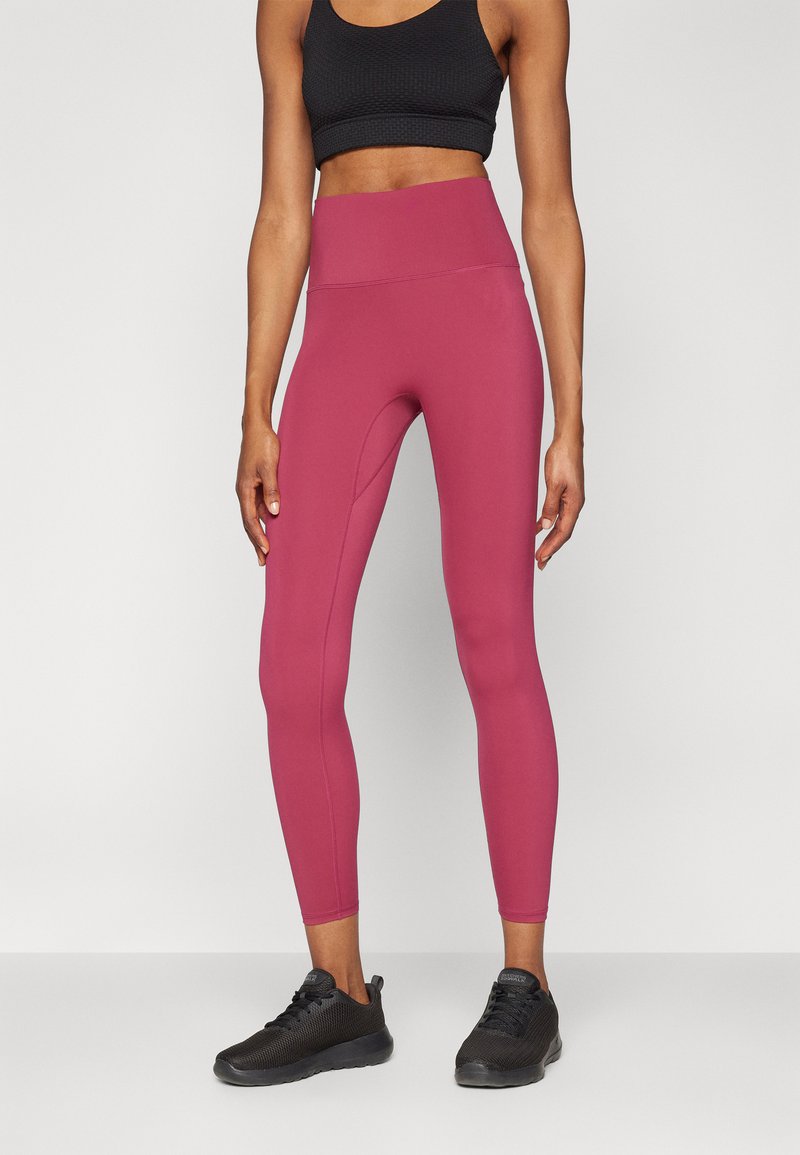 Leggings roses taille haute fabriqués dans un tissu extensible, avec une texture lisse et une silhouette ajustée, associés à des chaussures de sport noires.