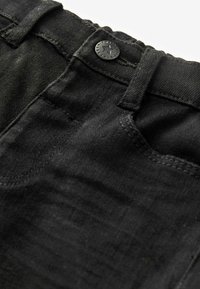 Los jeans de mezclilla negros cuentan con un cierre de cremallera, un botón y detalles de costura visibles. La tela tiene una textura suave con un ligero desgaste.
