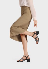 Sandali neri con tacco a blocco testurizzato e cinturini incrociati con borchie, indossati con una gonna chiara fino al ginocchio.