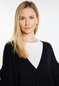 Fekete kötött kardigán mély V-nyakkivágással, középső gombbal és lazán álló ujjal. Fehér felsőre van húzva. Sima textúra.