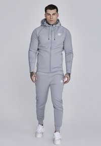 Šedá mikina na zips s kapucňou, obsahujúca logo a bočné zipsy, v kombinácii s rovnako farebnými teplákmi, oboje vyrobené z hladkej, elastickej látky.