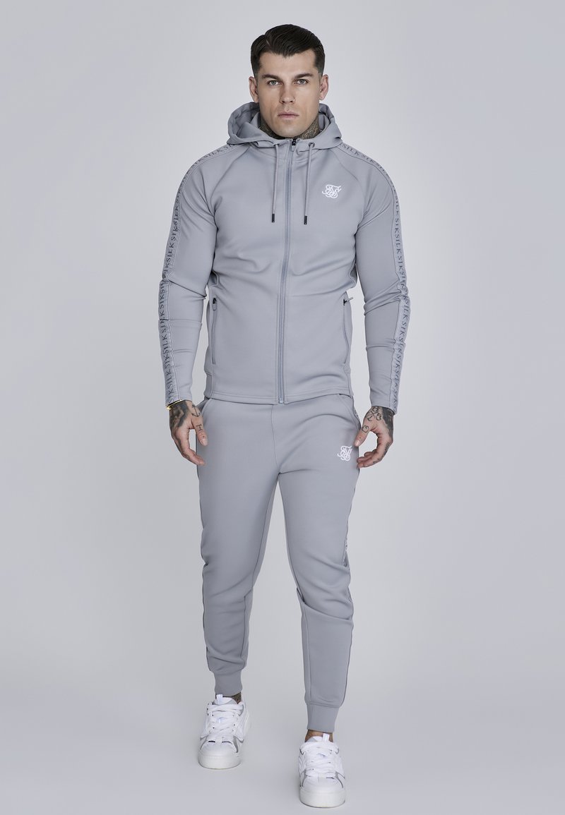 Grauer Reißverschluss-Hoodie mit Kapuze, versehen mit einem Logo und seitlichen Reißverschlüssen, kombiniert mit passender Jogginghose, beide aus glattem, elastischem Material gefertigt.
