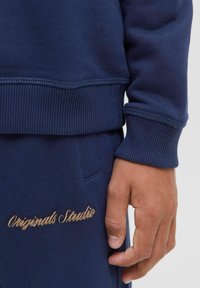 Navy blue sweatshirt and matching pants made of soft fabric, featuring ribbed cuffs and an embroidered "Originals Studio" logo in gold.