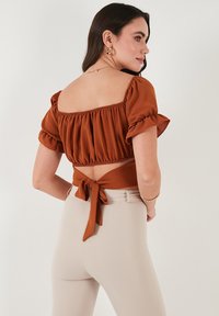 Rustfarvet crop top med puffede ærmer, elastisk udskæring og bundet detalje bagpå. Kombineret med lysebeige tætsiddende bukser.