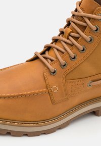 Bota de couro marrom com ilhós de metal, atacadores bege e sola robusta. Apresenta detalhes costurados e um bico reforçado para maior durabilidade.