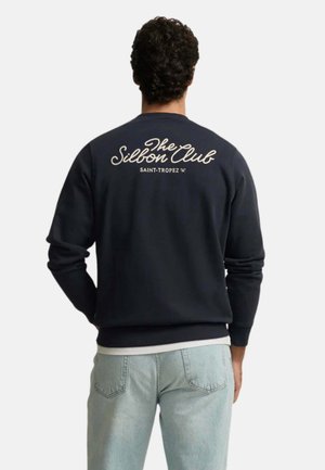 Hombre con sudadera azul marino con el texto "The Silbon Club Saint-Tropez" en la espalda, combinado con vaqueros azul claro, de espaldas.