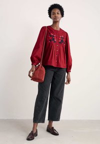 Červená vyšívaná blúzka s nafúknutými rukávmi, zapínanie na gombíky, skombinovaná s čiernymi voľnými nohavicami so zvýšeným pásom a hnedými mokasínami, drží malú kabelku.