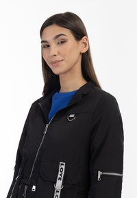 Giacca nera con zip, caratterizzata da un colletto alto, due tasche anteriori e un nastro decorativo con la scritta "LOVE". Indossata sopra un top blu.
