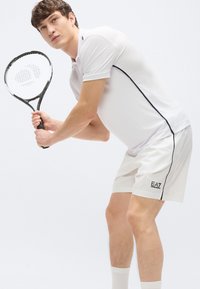 Biely polokošeľ s čiernymi detailmi v kombinácii s bielymi šortkami. Model drží čiernu tenisovú raketu a stojí v dynamickej póze.