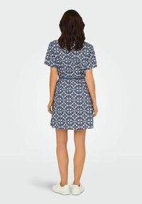 Mornarsko plava haljina s kratkim rukavima, ukrašena bijelim geometrijskim oblicima, sužena u struku i s raširenom suknjom, uparena s bijelim tenisicama.