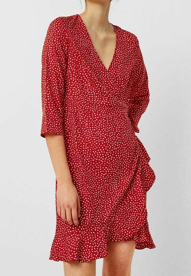 Femme portant une robe portefeuille rouge à pois blancs, manches trois-quarts et ourlet à volants sur un fond clair uni.
