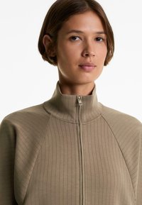 Giacca zip-up beige con texture rigata e colletto alto. Caratterizzata da un motivo a righe verticali e dettagli di zip argentata sulla parte anteriore.