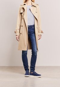 Beiger Trenchcoat mit doppelreihigen Knöpfen, einem taillierten Gürtel und Schulterklappen, getragen über einem gestreiften Hemd und blauen Jeans.