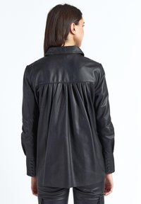 Sort læderjakke med plisseret ryg, krave og manchetter med knapper. Glat tekstur og skræddersyet pasform.