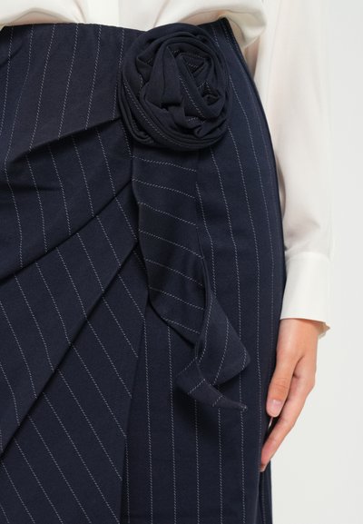 Marineblauwe krijtstreep rok met plooien en een stoffen rozen-detail op de taille, gedragen met een witte blouse met lange mouwen.