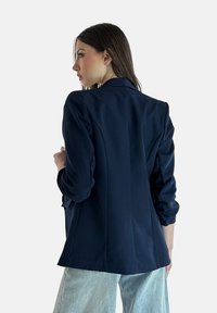 Blazer blu navy realizzato in tessuto liscio, con una vestibilità sagomata, revers a taglio e maniche lunghe, visto da dietro.
