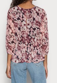 Donna che indossa una blusa floreale rosa e bordeaux con maniche a 3/4 e chiusura a bottone sul retro a forma di chiave, abbinata a jeans in denim blu chiaro.