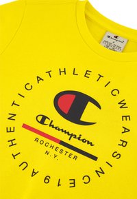 Camisa de algodón amarilla con un diseño de logo circular que incluye el texto "Champion" en negro, acentos rojos y la fecha de establecimiento "1919."