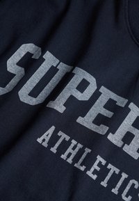 Marineblauwe stof met grote grijze tekst "SUPER" en kleinere tekst "ATHLETIC" gedrukt over het oppervlak.