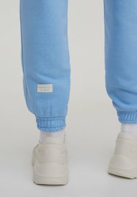 Pantalons de survêtement bleu clair avec des poignets élastiques, arborant une petite étiquette blanche avec l'inscription "JOURNÉES SMOOTHIE." Associés à des baskets blanches et des chaussettes blanches.