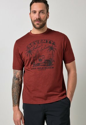 Mand med skæg og tatoveret arm iført en rust-rød t-shirt med teksten "Seventy Wheels & Waves Five Mile Beach Australia" og mørke shorts.